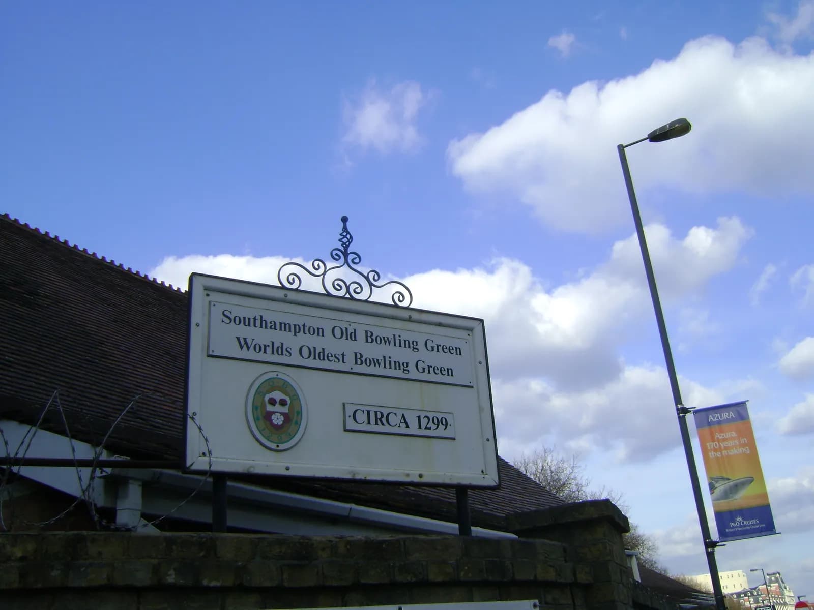 A white sign with ornate scrollwork at the top proudly announces "Southampton Old Bowling Green World's Oldest Bowling Green CIRCA 1299." The sign is set against a blue sky with fluffy white clouds, with the roofline of a building and part of a street light visible.