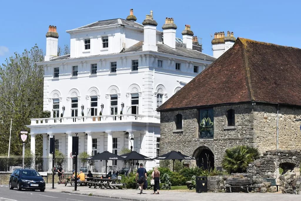 A historic white building with columns and a balcony stands next to a stone building with a terracotta roof, with people seated at outdoor tables and walking on the street.