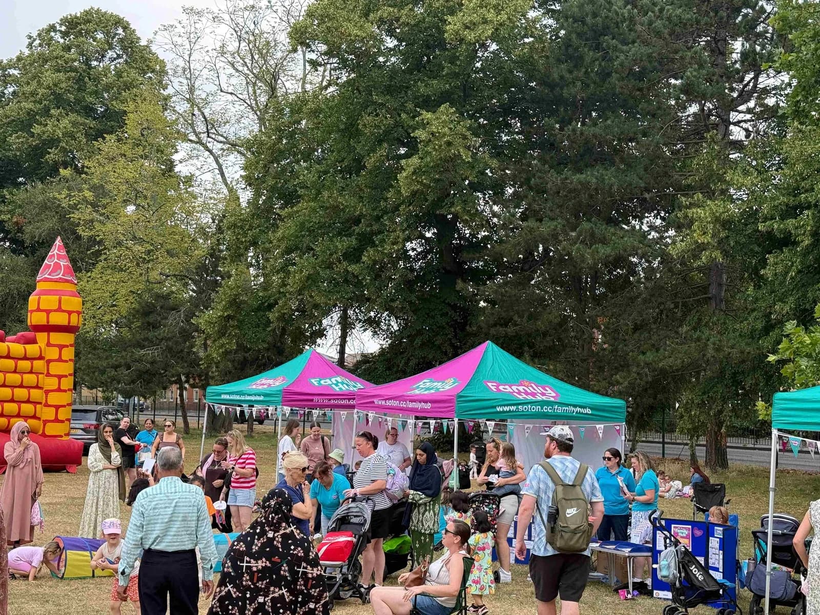 A diverse crowd of people gathered under two large green and pink tents for a 'Family Hub' event, with a bouncy castle to the left and trees in the background.