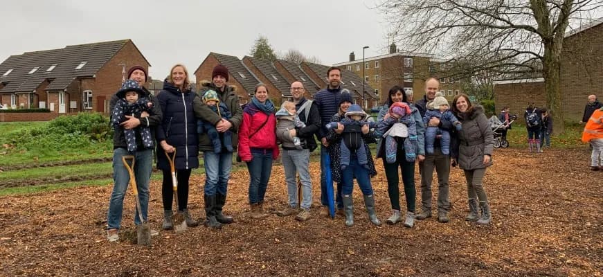 A group of parents and their young children stand together, some holding shovels, on a patch of dirt and woodchips in front of suburban houses on a cloudy day.