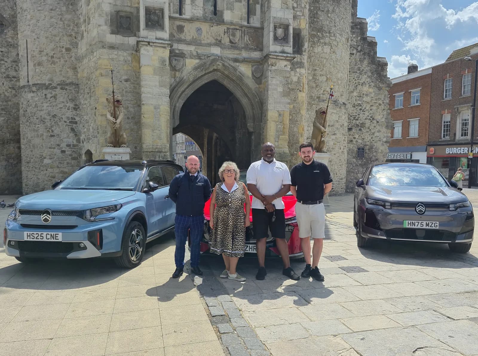 Four people standing outdoors in front of two cars and a stone archway, with the two men on the right standing in front of a red car.