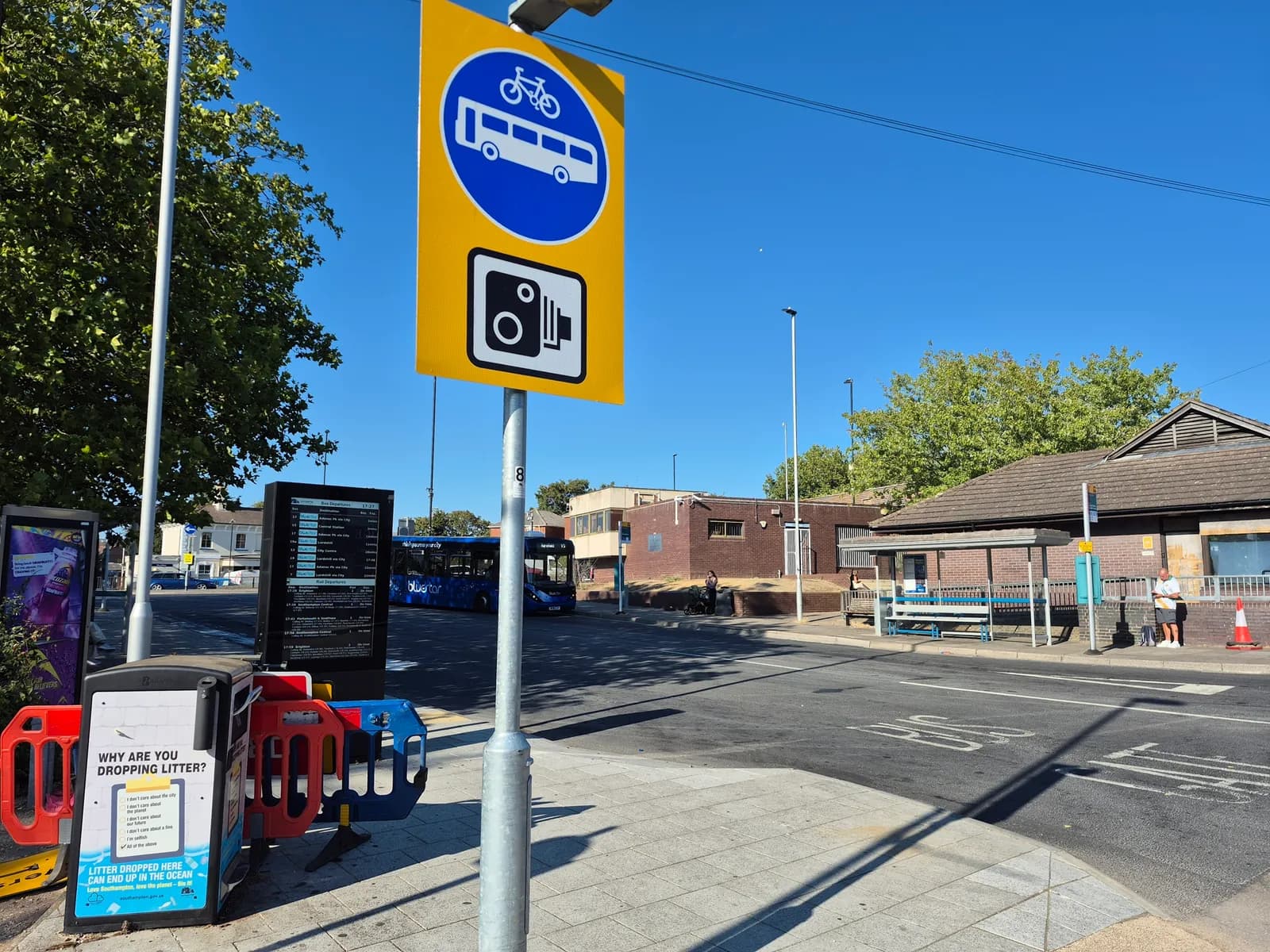 A yellow sign with blue symbols for bus, bicycle, and a camera on a pole beside a road, with bus stop benches and buildings in the background under a clear sky.