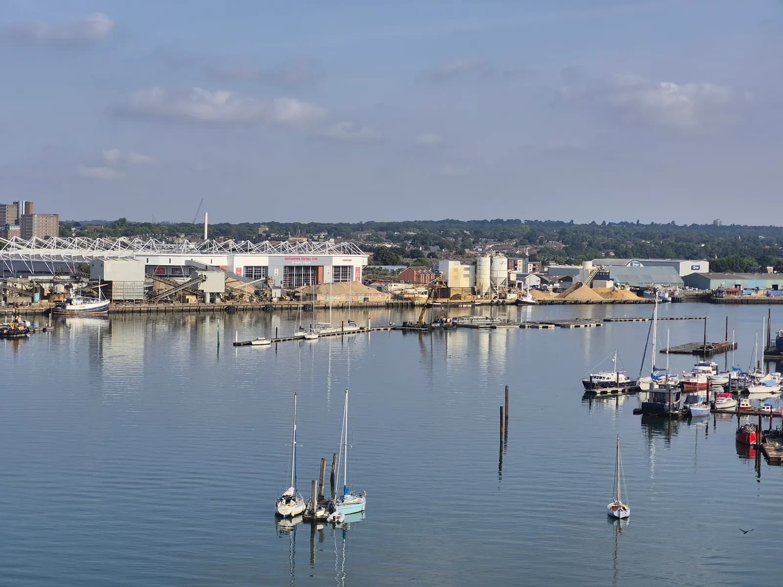 A wide shot of a bustling port with multiple boats docked in the calm blue water and industrial buildings lining the distant shore under a partially cloudy sky.