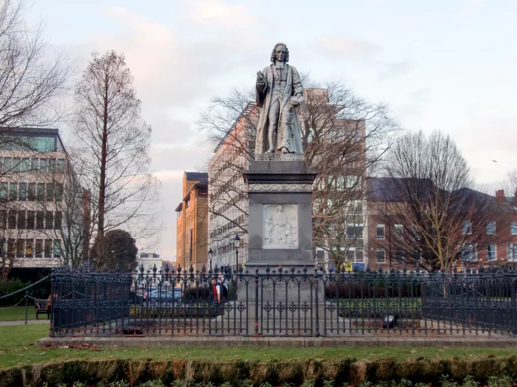 A stone statue of a man in religious attire stands on a pedestal surrounded by a black wrought iron fence, with buildings and bare trees in the background under a cloudy sky.