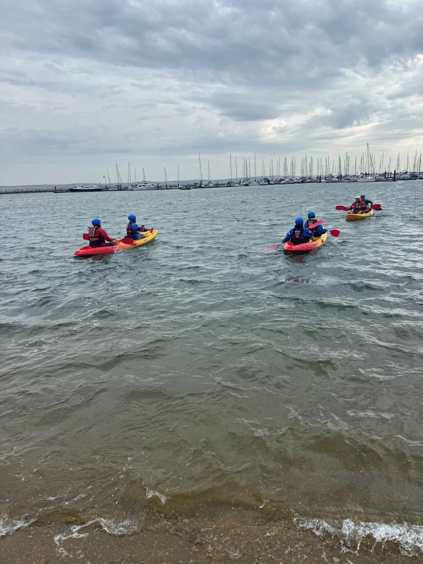 A group of people kayaking on a body of water with a marina visible in the background under an overcast sky.
